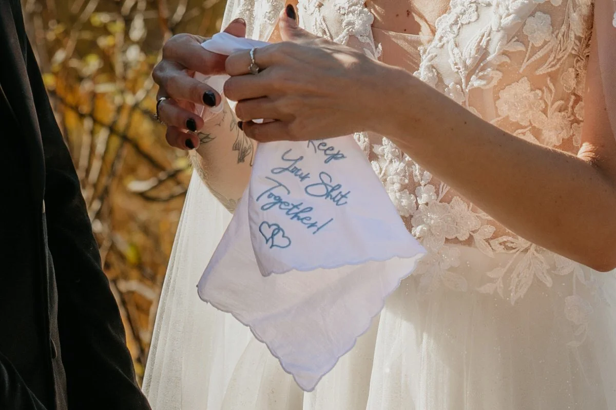 Close-up of bride holding a white embroidered handkerchief that reads “Keep Your Shit Together” with a small heart, sunlight highlighting lace details on her wedding dress and her rings.
