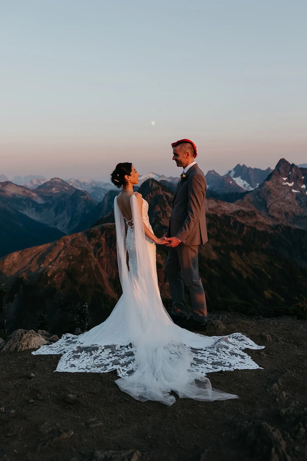 Newly married couple standing on a mountaintop at sunrise overlooking rugged North Cascades peaks.