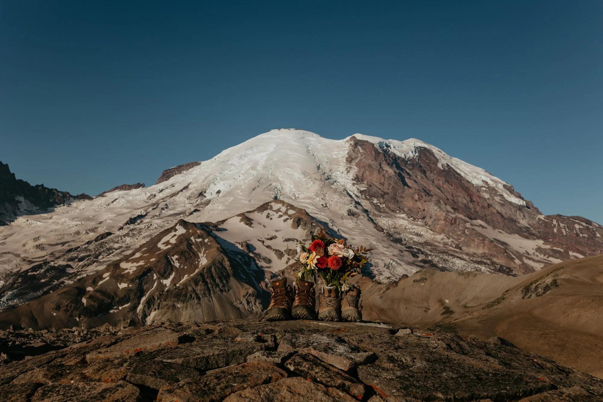 Hiking boots and elopement florals placed on a rocky overlook with a snow-capped mountain dominating the horizon