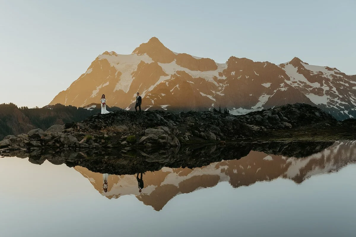 Wide landscape view of couple standing on a rocky ridge with Mount Shuksan and its reflection in an alpine tarn.jpg