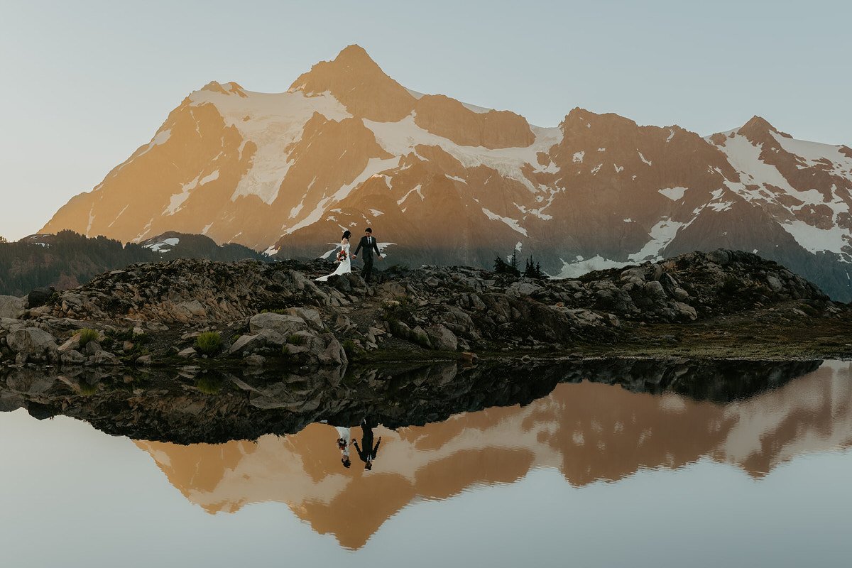 Bride and groom walking hand in hand across a rocky overlook with Mount Shuksan glowing in early morning light.jpg