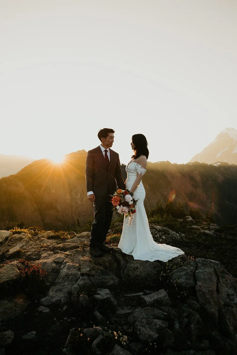 Bride and groom standing on a rocky cliff at sunrise with sun rays breaking over the ridge behind them.jpg
