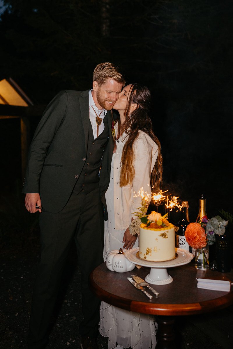 Couple kissing beside their wedding cake and sparklers during an evening reception