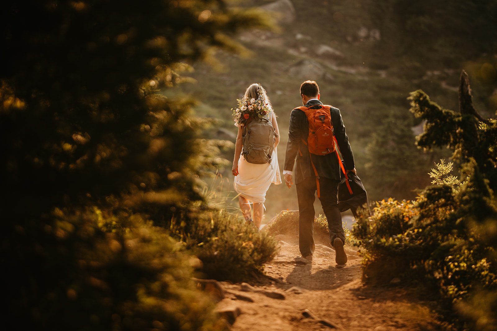 Couple walking hand in hand down a sunlit dusty trail after their ceremony, backpacks in tow