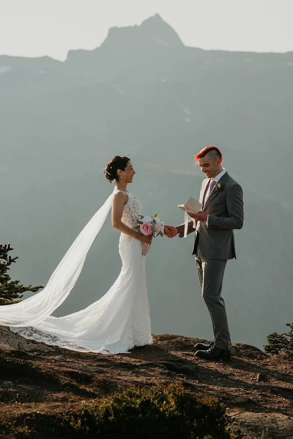 A couple exchanges vows during an intimate mountaintop elopement, with the bride’s long veil flowing in the wind as the groom reads from a vow book against a misty mountain backdrop.