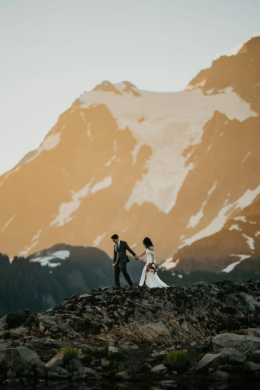 Eloping couple walking hand in hand along a rocky ridge with dramatic golden light illuminating snow covered mountain peaks behind them