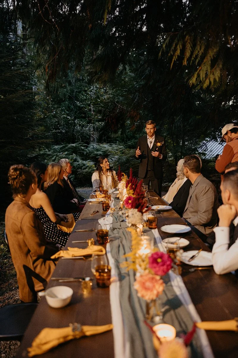 Groom giving a heartfelt speech at a candlelit outdoor reception table surrounded by guests in a forest setting after a Washington elopement ceremony.
