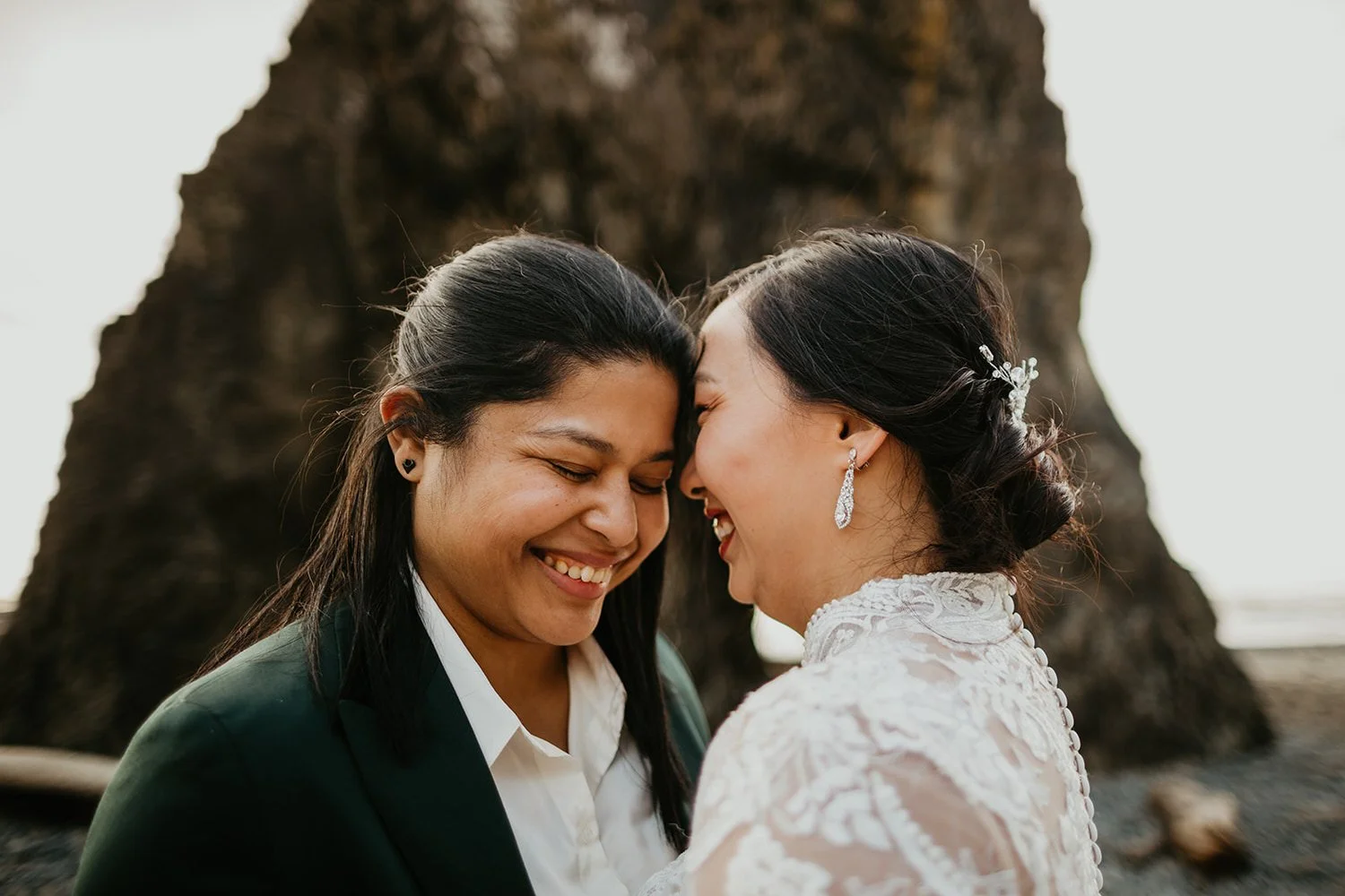 Close-up of two brides smiling and touching foreheads on a rocky beach with a large sea stack in the background.