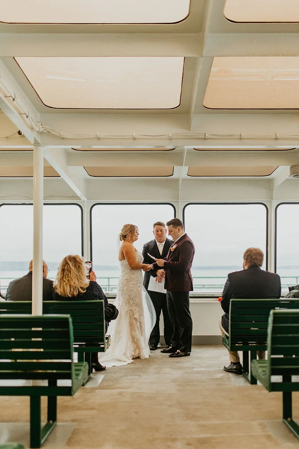 A couple exchanges vows during an intimate ferry elopement ceremony, standing between green benches as a small group of guests watches through large windows overlooking the water.
