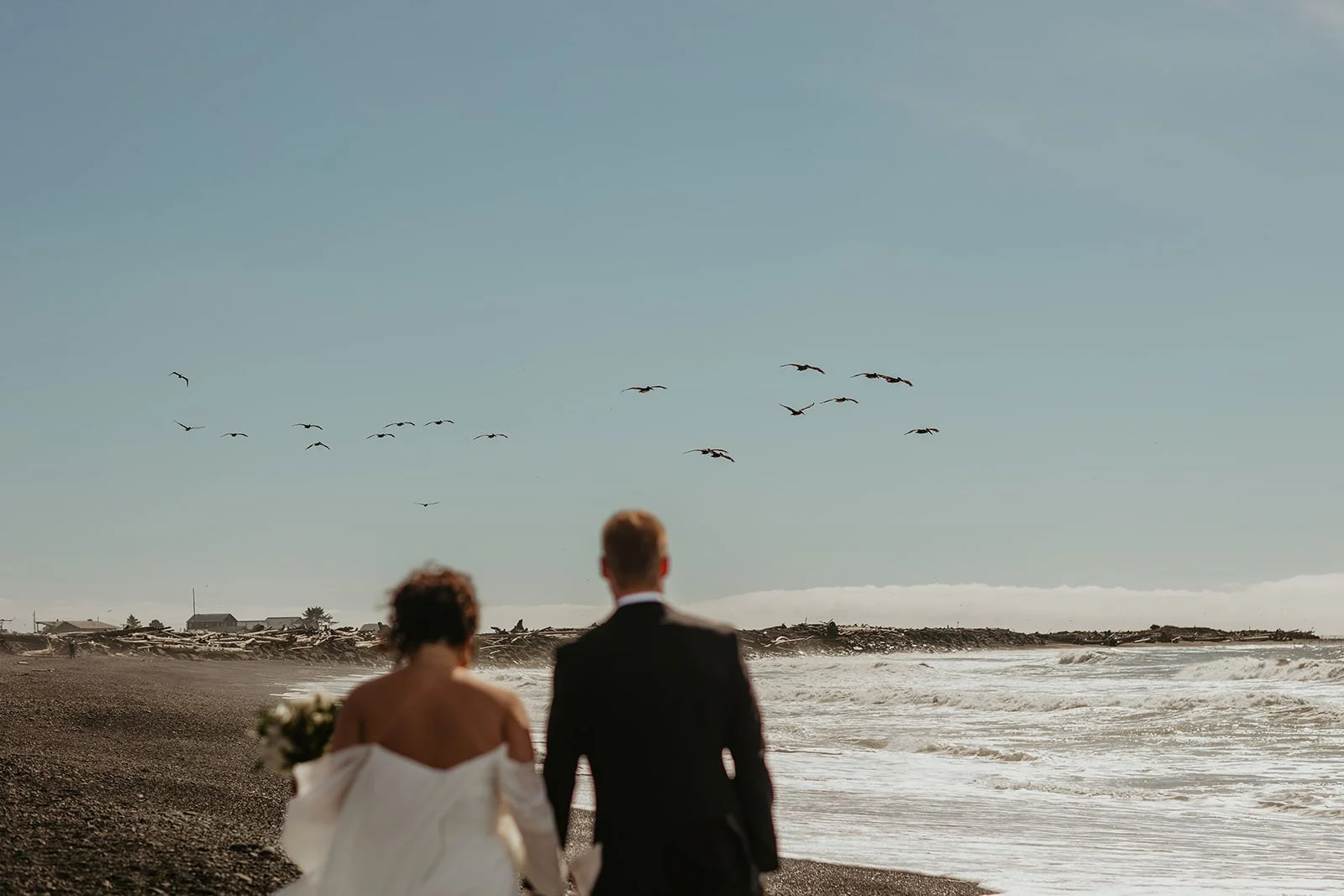 Elopement couple walking along a windswept beach as birds fly overhead, waves crashing nearby.