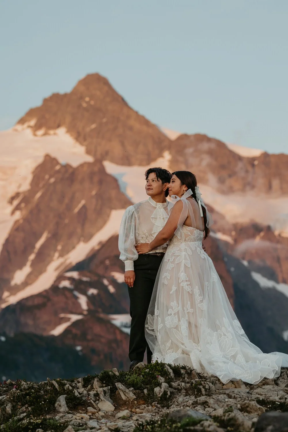Two brides standing together on a rocky alpine ridge at golden hour, wearing lace wedding dresses with snow-covered mountain peaks glowing behind them.