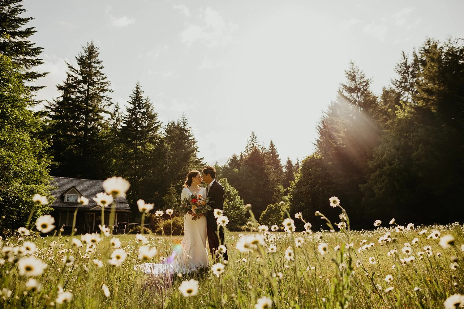 Couple embracing in a sunlit meadow filled with wildflowers, with a historic cabin and dense forest in the background.