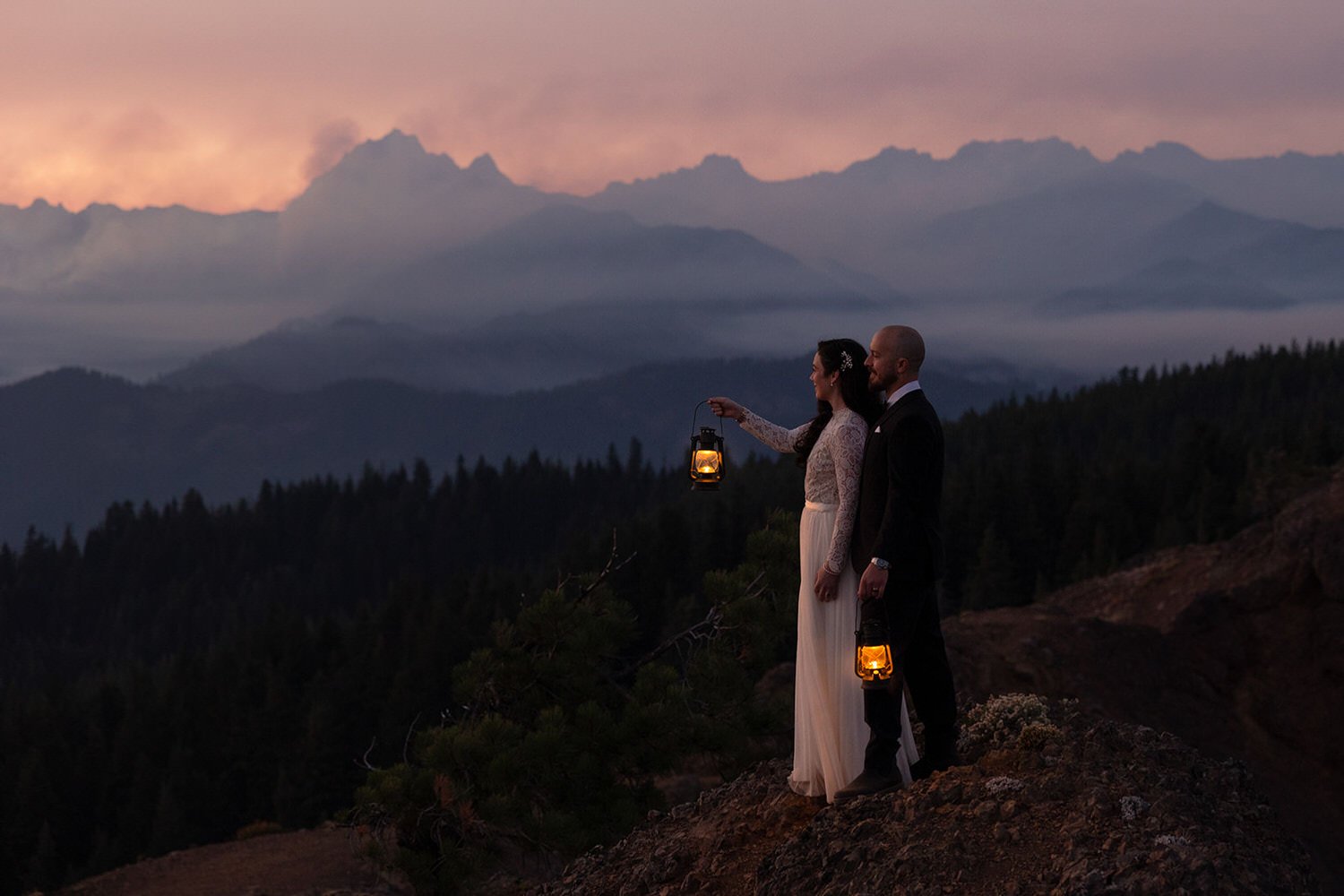 Elopement couple holding lanterns at dusk on a mountain overlook, surrounded by layered ridgelines and soft evening light.
