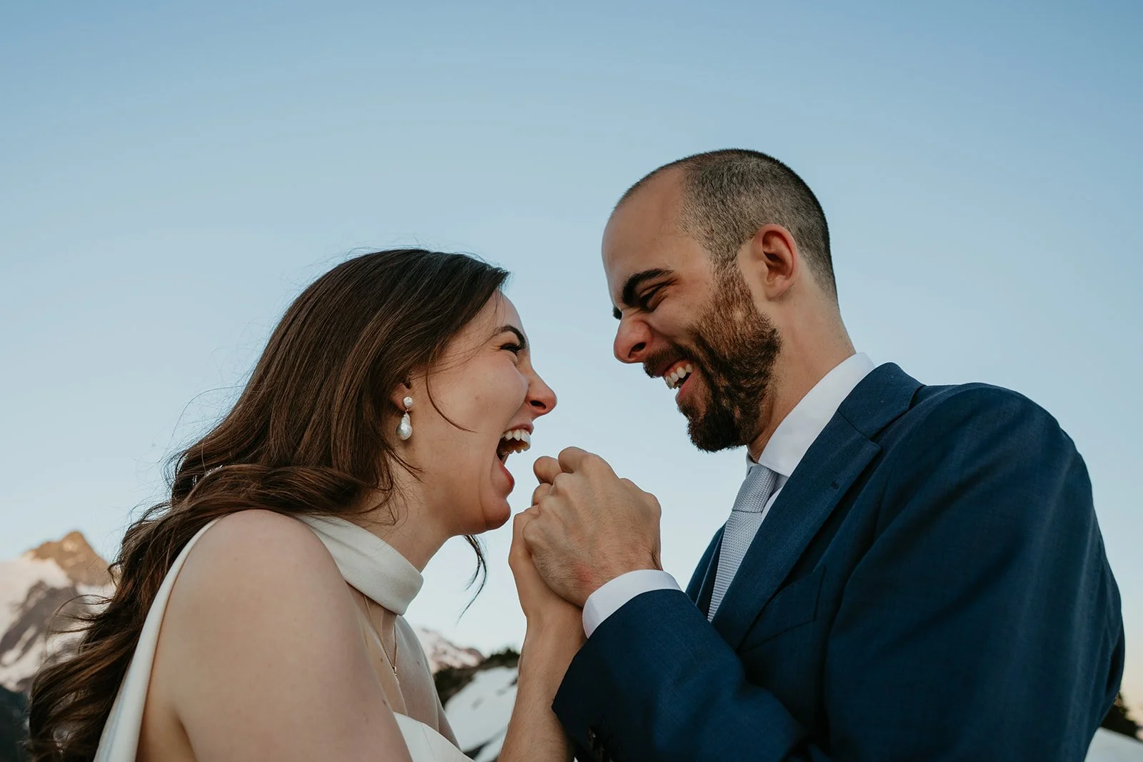 A smiling couple, a woman with long brown hair and pearl earrings and a man with a beard and a suit, are face to face outdoors with mountain snow in the background, appearing joyful and close.