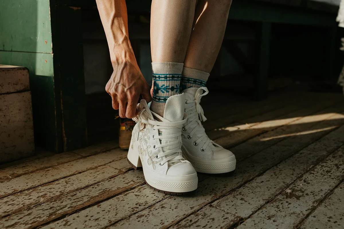 Bride lacing up white lace-up ceremony boots over knit socks inside a rustic cabin before an outdoor elopement.