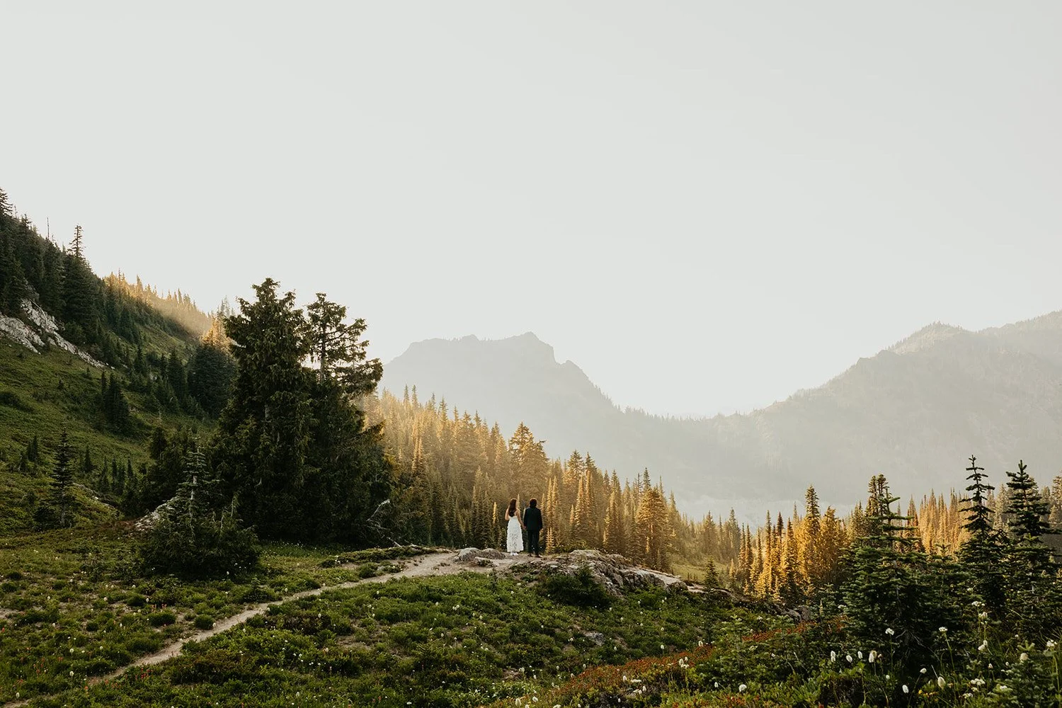 A couple stands on a rocky overlook surrounded by alpine meadows and evergreen trees, facing distant mountain ridgelines glowing in soft evening light.