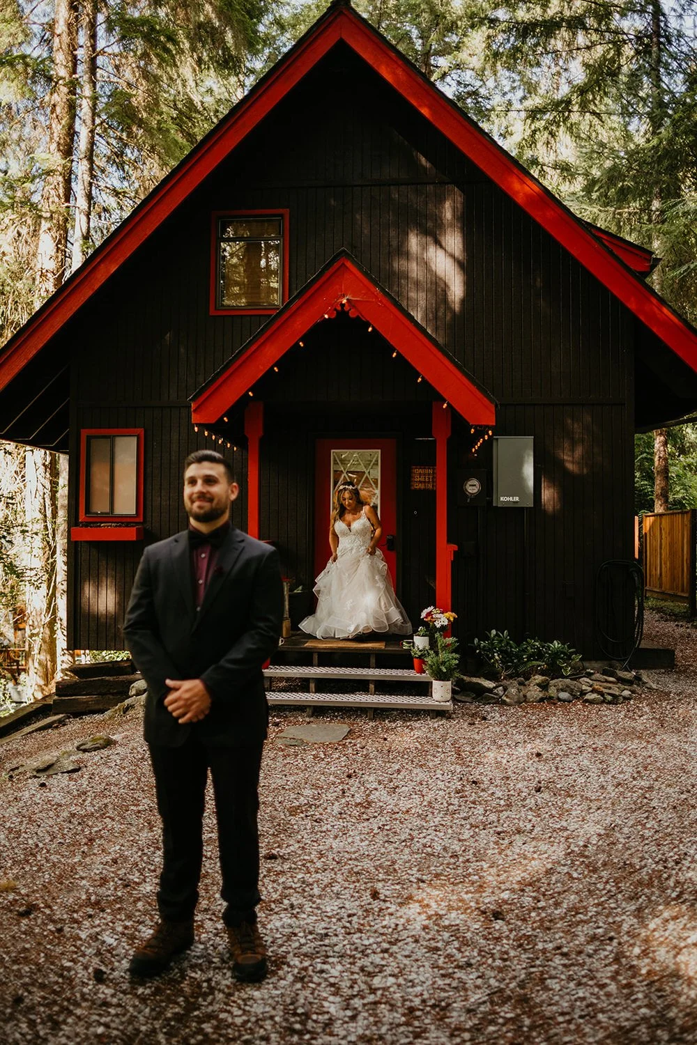 Groom standing at the entrance of a red cabin door with flowers, bride beside him, during a Washington Airbnb elopement