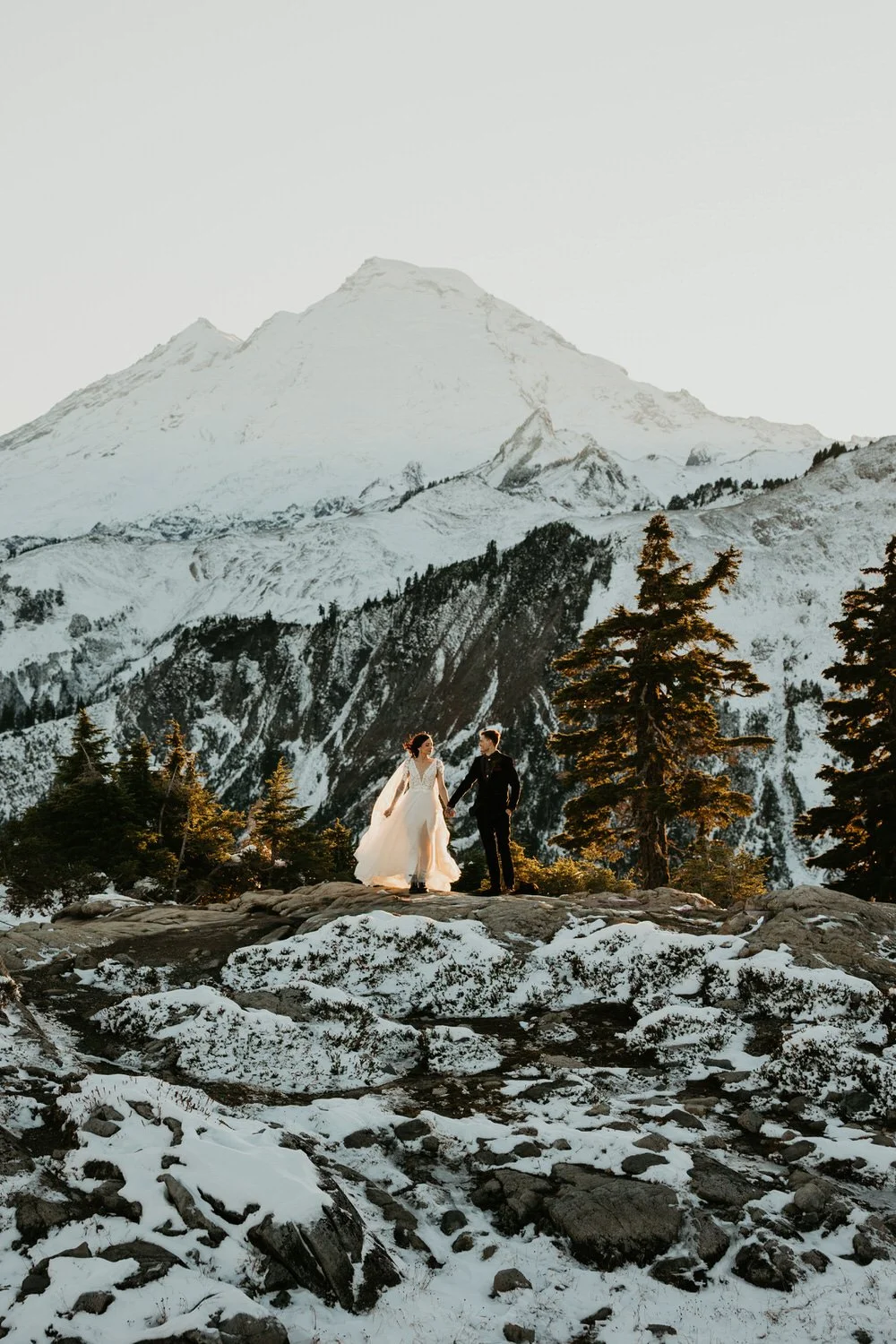 Couple walking hand in hand across a rocky ridge with snow-covered Mount Baker rising behind them