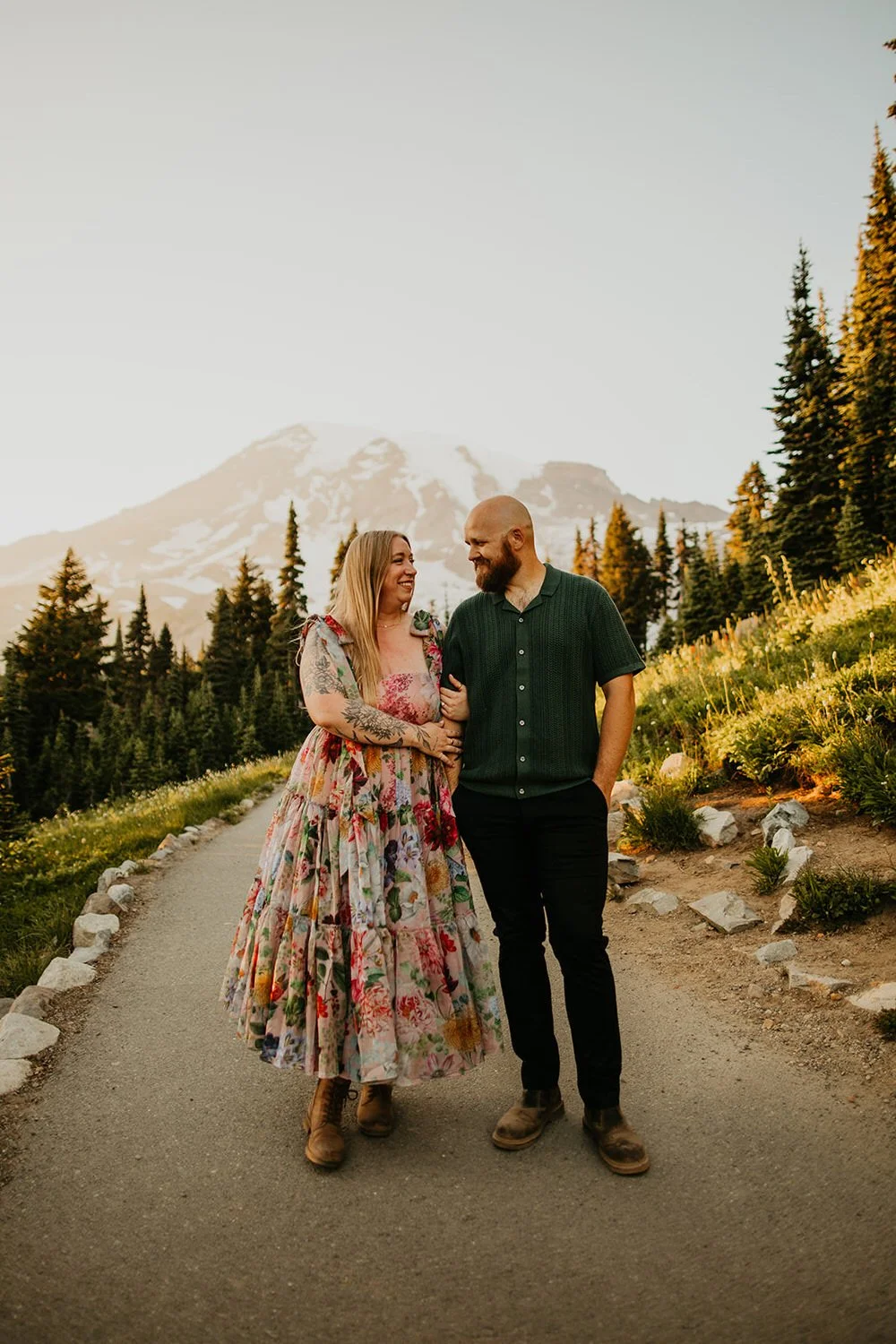 A couple walks arm in arm along a paved trail lined with wildflowers and evergreen trees, with Mount Rainier rising softly in the background.