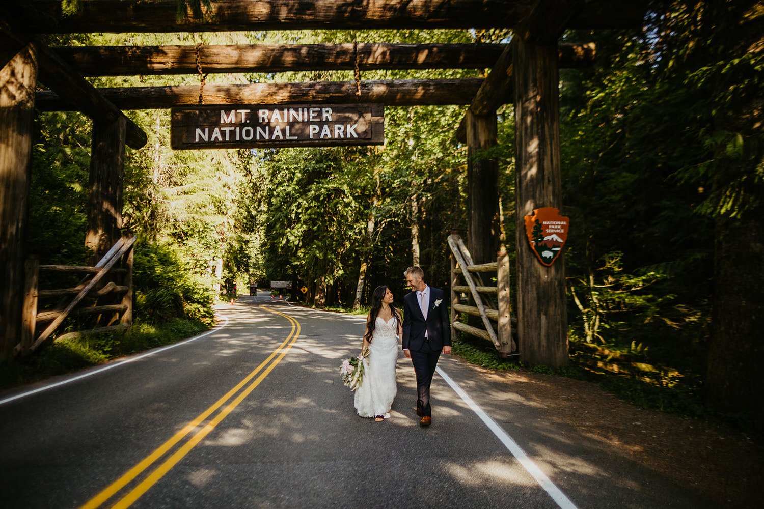 A couple in wedding attire walks hand in hand beneath the Mount Rainier National Park entrance sign on a quiet forest road.