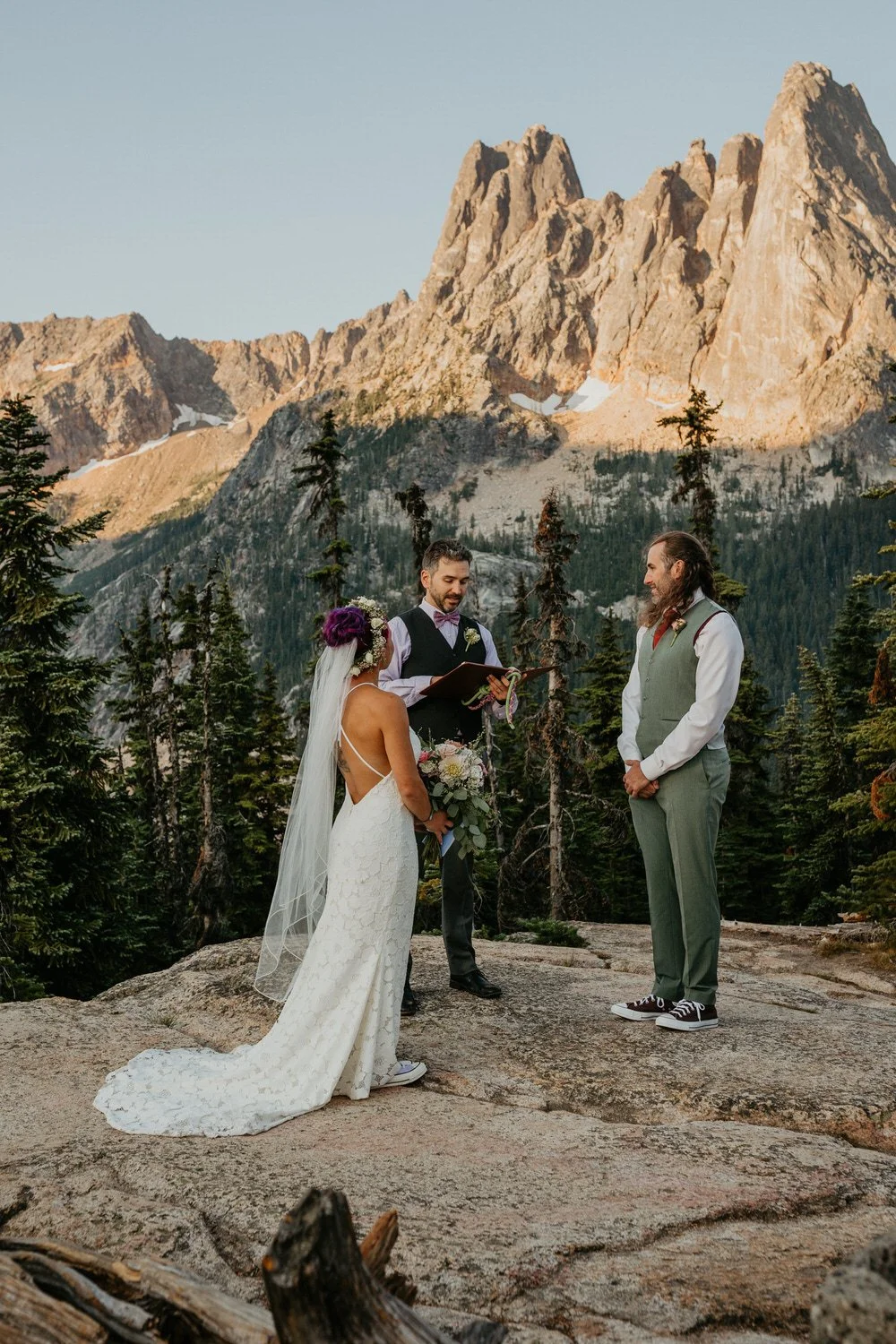 A small elopement ceremony takes place on a rocky alpine overlook, surrounded by evergreen trees, with jagged North Cascades peaks rising dramatically in the background under soft evening light.