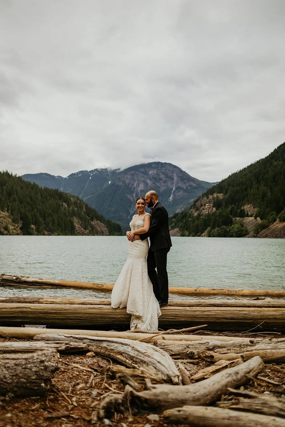 Couple embracing on driftwood along the turquoise Diablo Lake in the North Cascades, surrounded by forested mountains