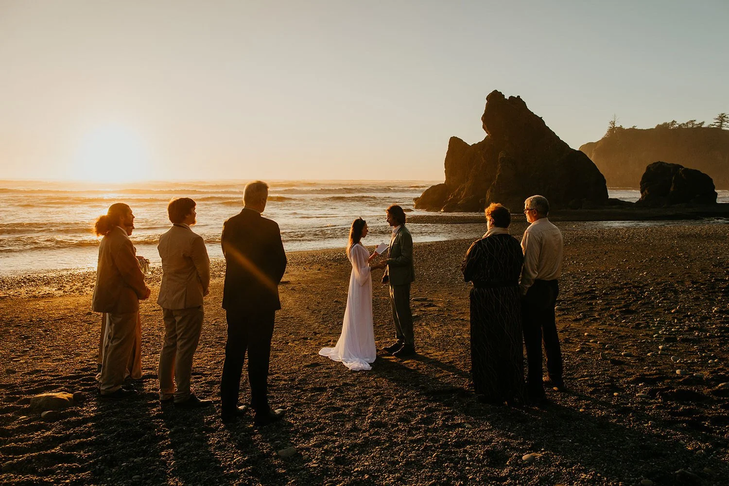 Small beach elopement ceremony at sunset, with a couple exchanging vows in front of close family as waves roll in and sea stacks rise behind them.