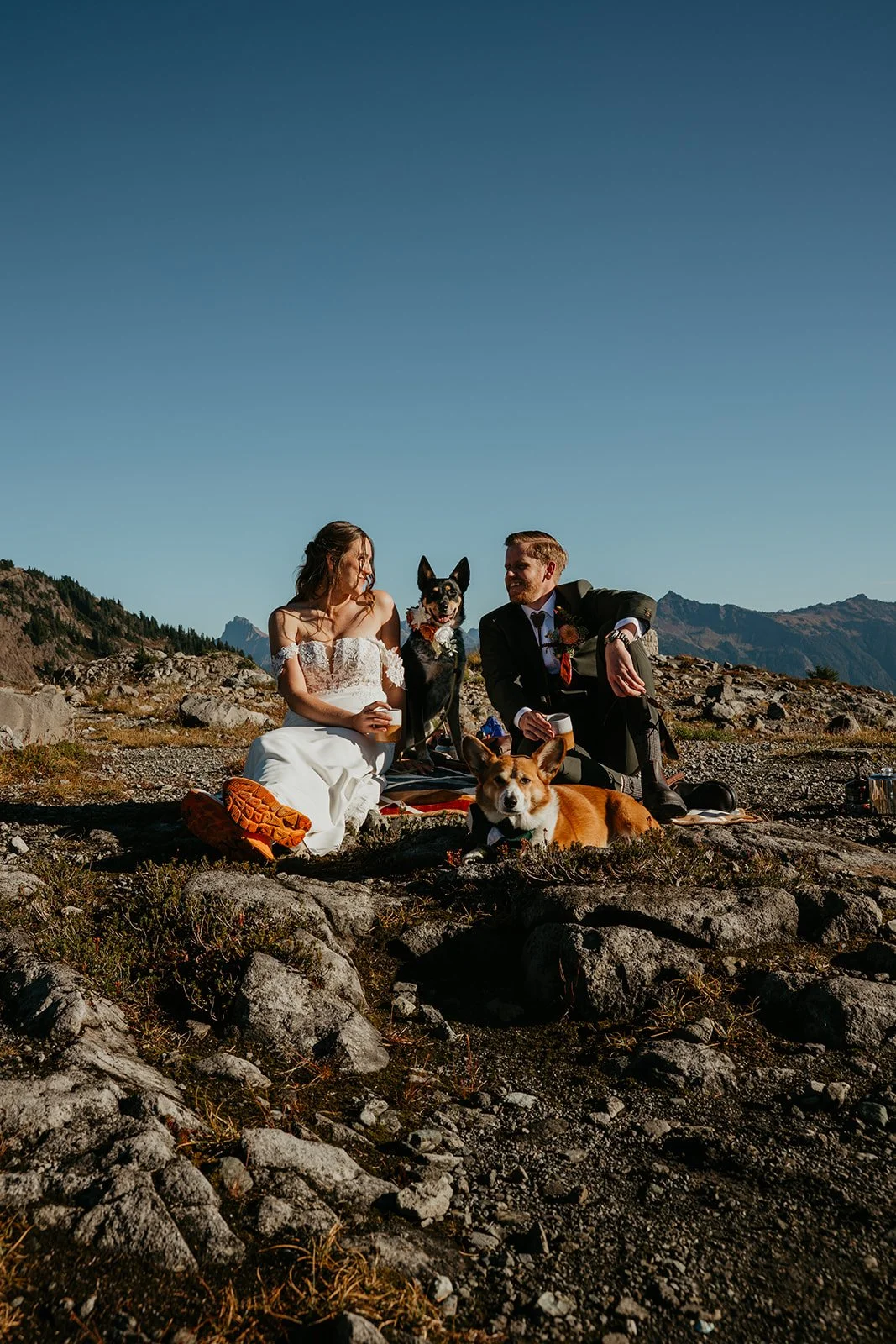 An eloping couple drinks their morning coffee on the trail with their two dogs sitting with them
