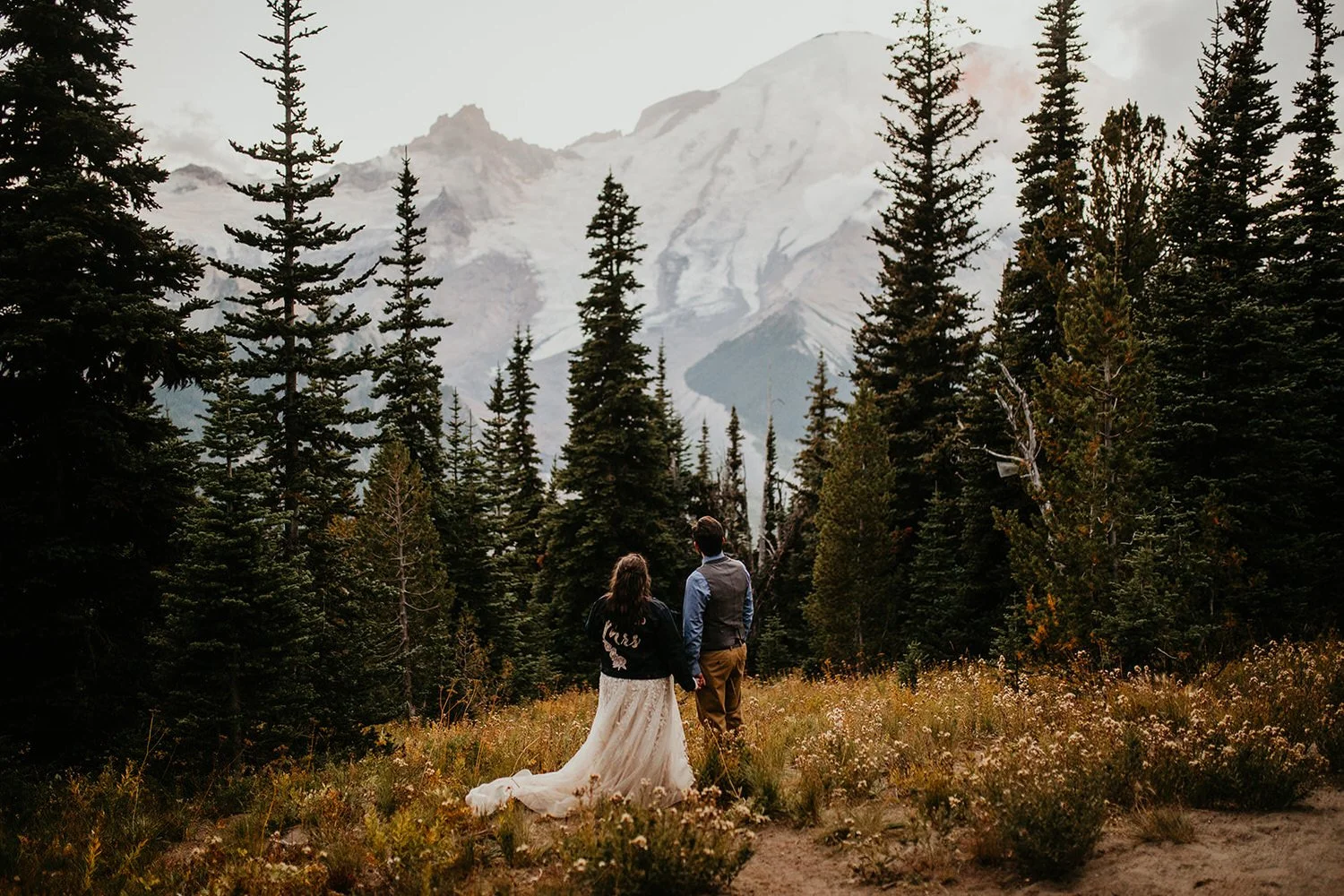 A couple in wedding attire stands in a wildflower meadow surrounded by evergreen trees, looking toward Mount Rainier’s snow-covered peak in the distance.