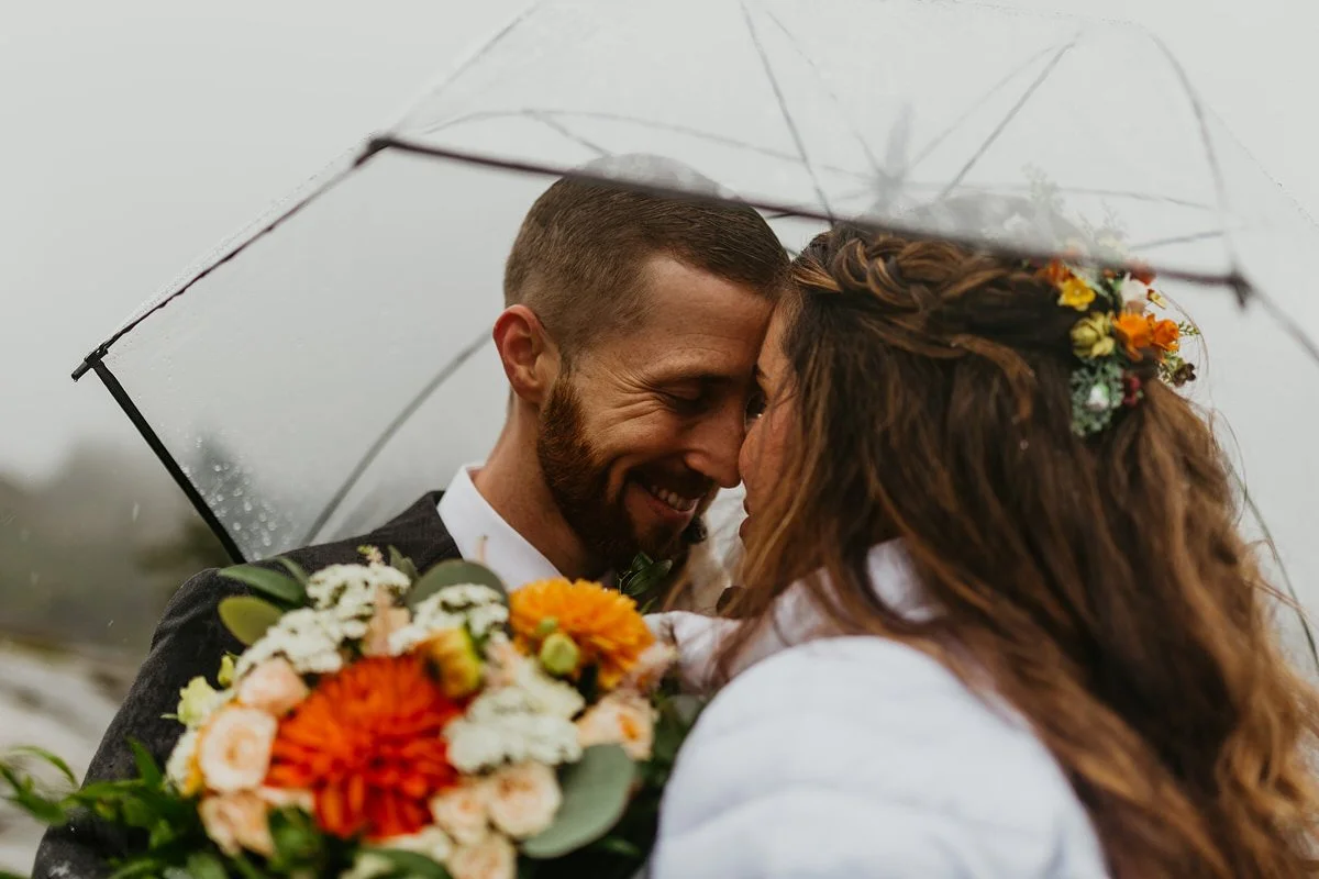 Newlyweds leaning their foreheads together beneath a transparent umbrella as rain falls around them, bouquet in the foreground.