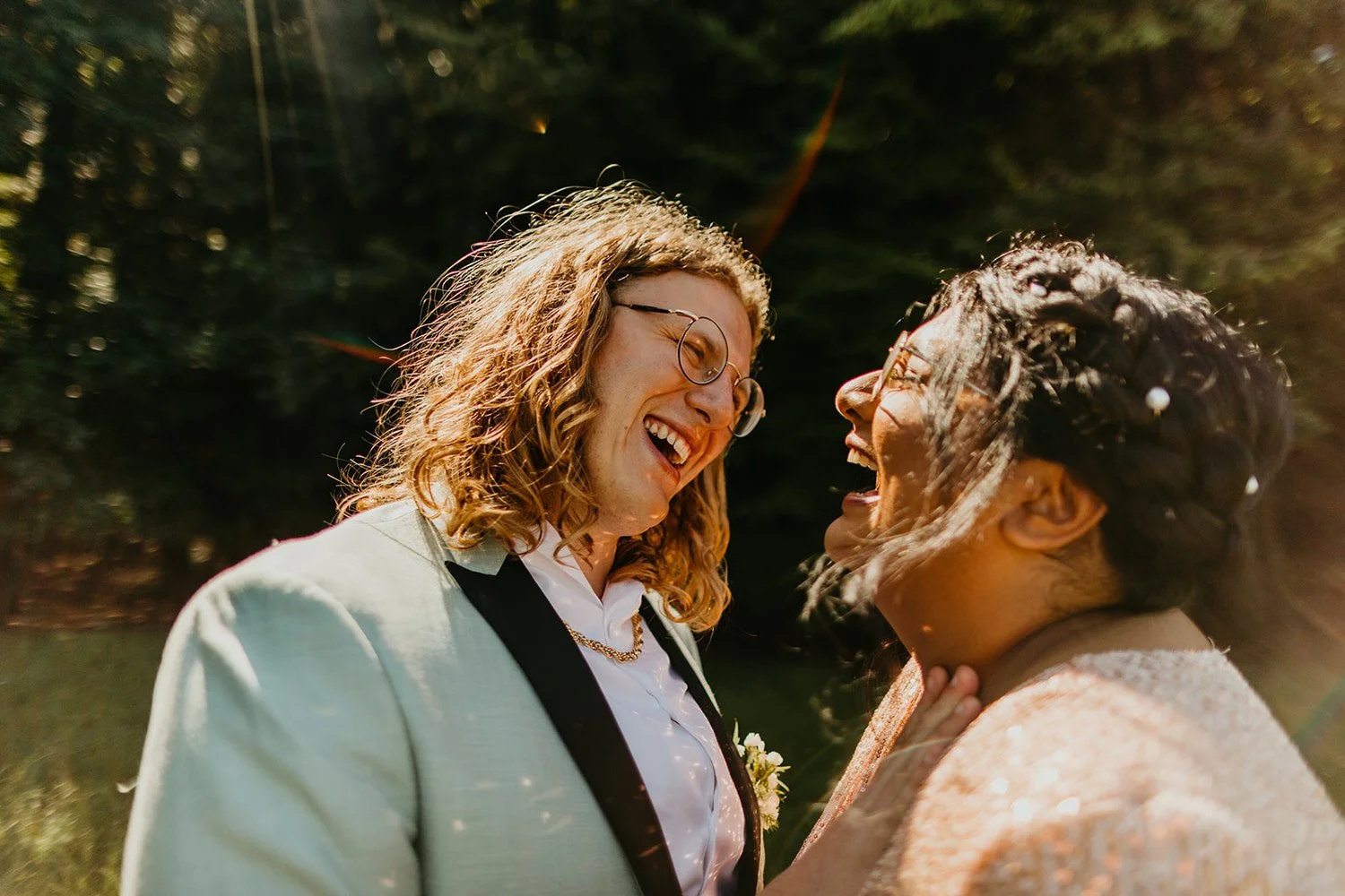Two newlyweds laughing together during golden hour, wearing wedding attire and standing outdoors with sunlight filtering through trees.