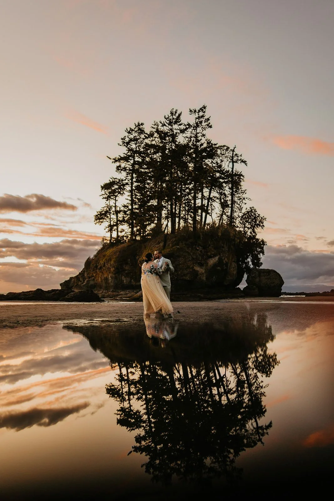 Eloping couple standing together at low tide as their reflection mirrors a small tree-covered sea stack against a colorful sunset sky.