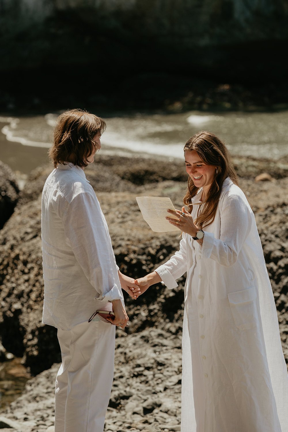 Queer couple holding hands during a private elopement ceremony on a rocky shoreline near the ocean.