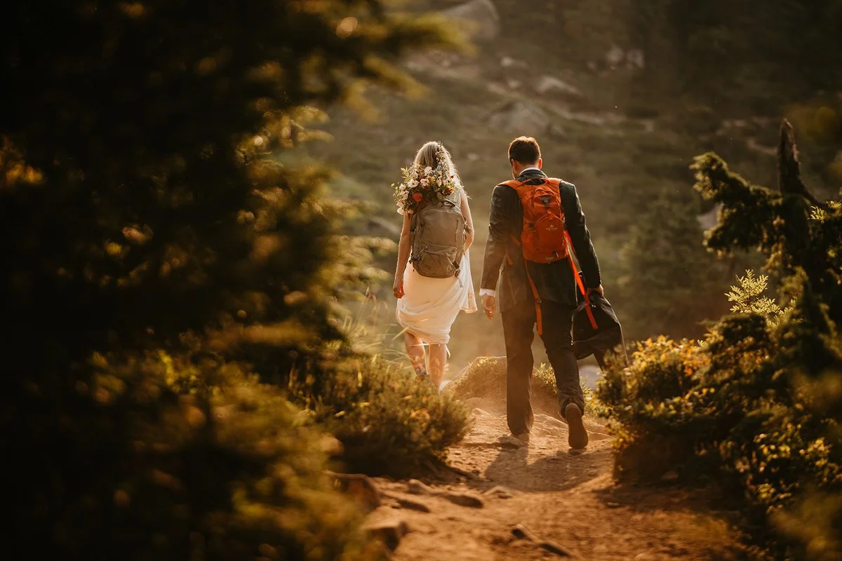 Bride and groom walking down a sunlit forest trail with backpacks during their Washington hiking elopement adventure.