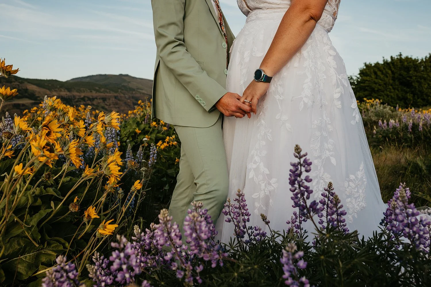 Close up of an eloping couple holding hands in a wildflower meadow with lupine and yellow blooms surrounding a wedding dress and green suit
