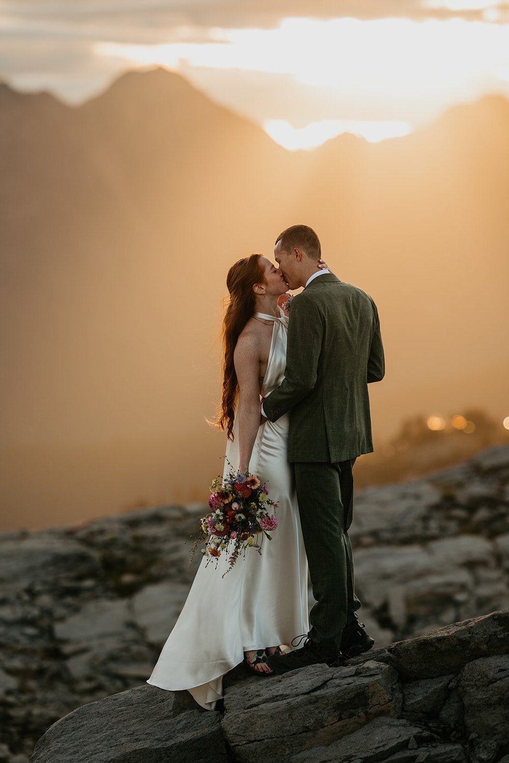 Couple sharing a kiss on a rocky mountain overlook at sunset, warm golden light and hazy peaks in the background