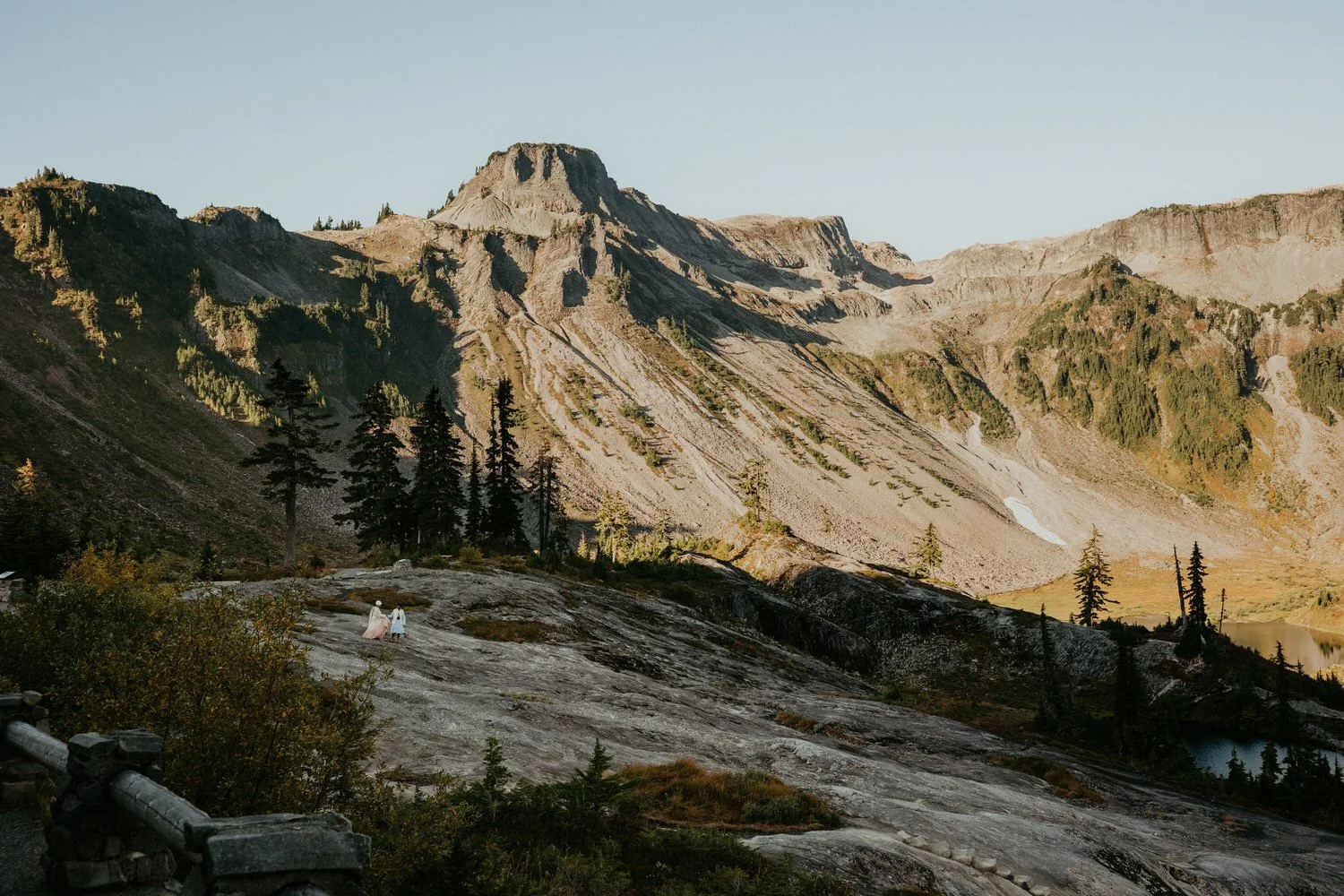 Wide view of two figures walking across granite slabs in the Chain Lakes basin at evening light