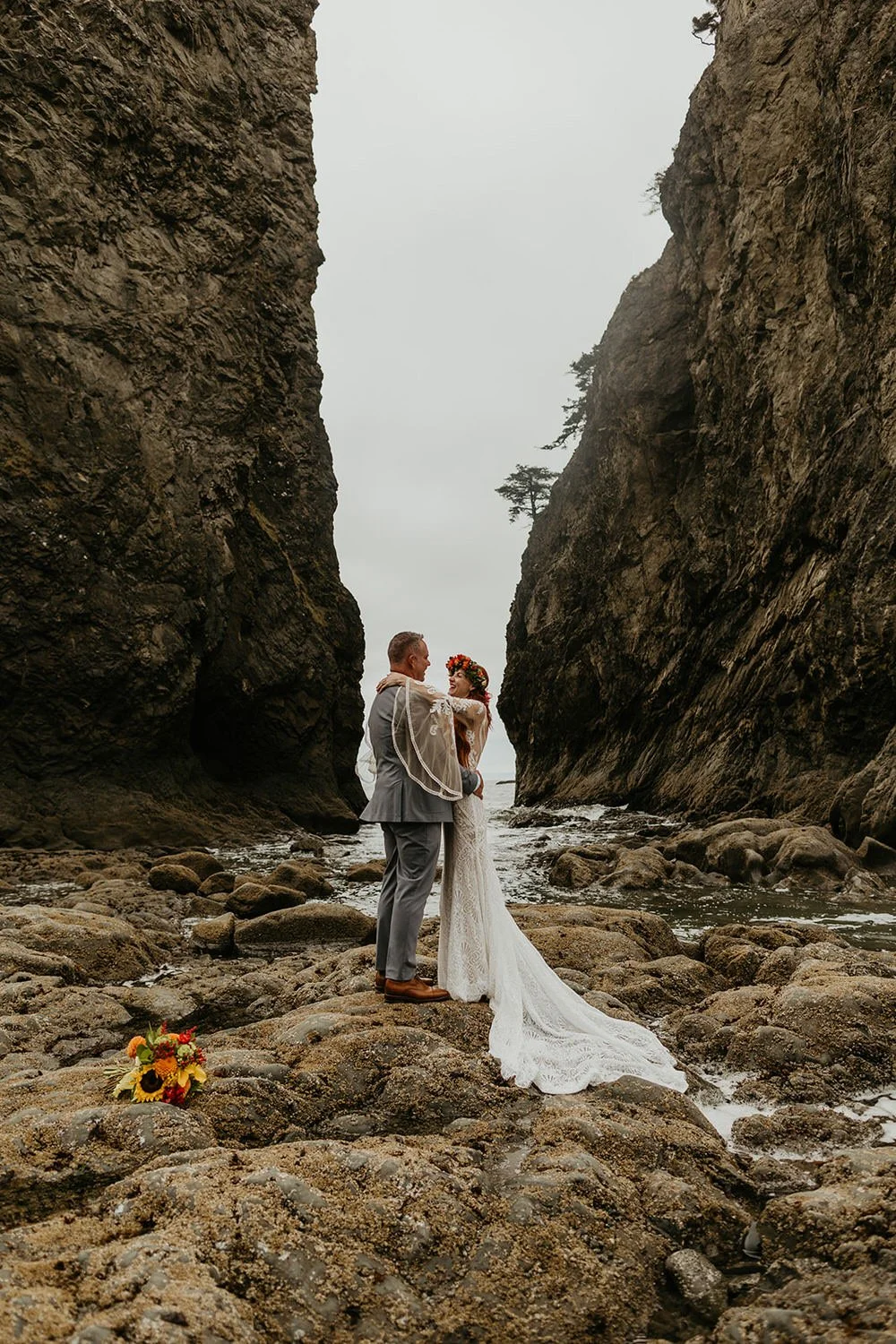 Eloping couple standing on a rocky beach between tall coastal cliffs, embracing beside tide pools with the ocean behind them