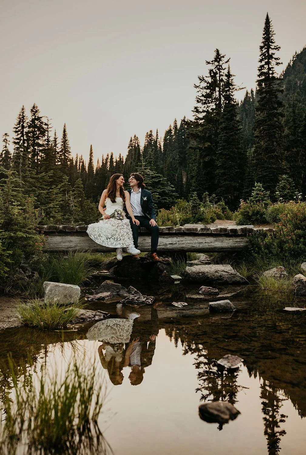 A couple sits together on a fallen log beside a shallow creek, surrounded by evergreen trees reflected in the still water.
