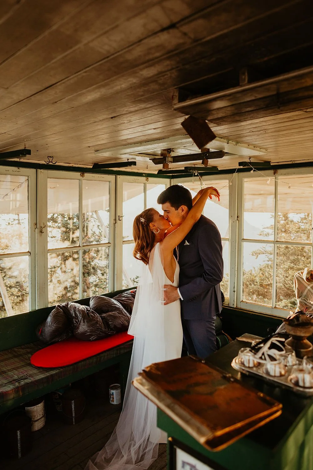 A kissing couple stands in a rustic fire lookout after their elopement ceremony, surrounded by large windows and sunlight
