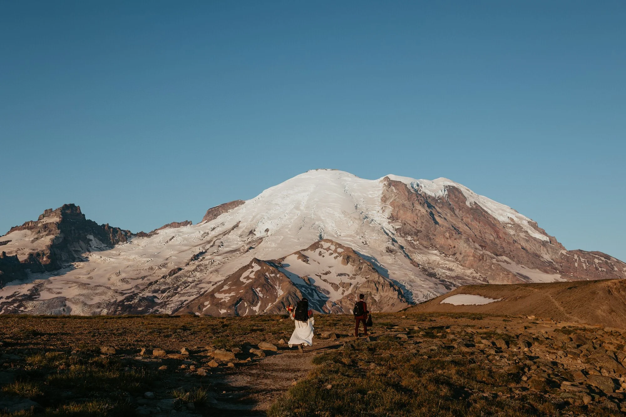 A couple hikes across a rocky alpine landscape toward Mount Rainier, with the snow-covered mountain dominating the background.
