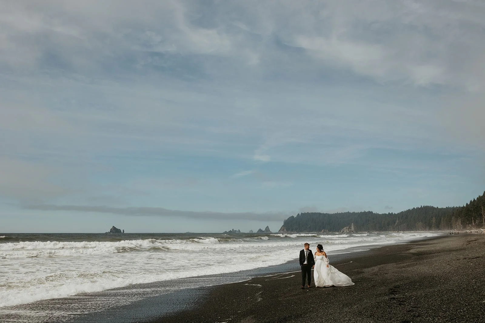 Eloping couple walking along a windswept black sand beach with waves crashing nearby and sea stacks visible in the distance.