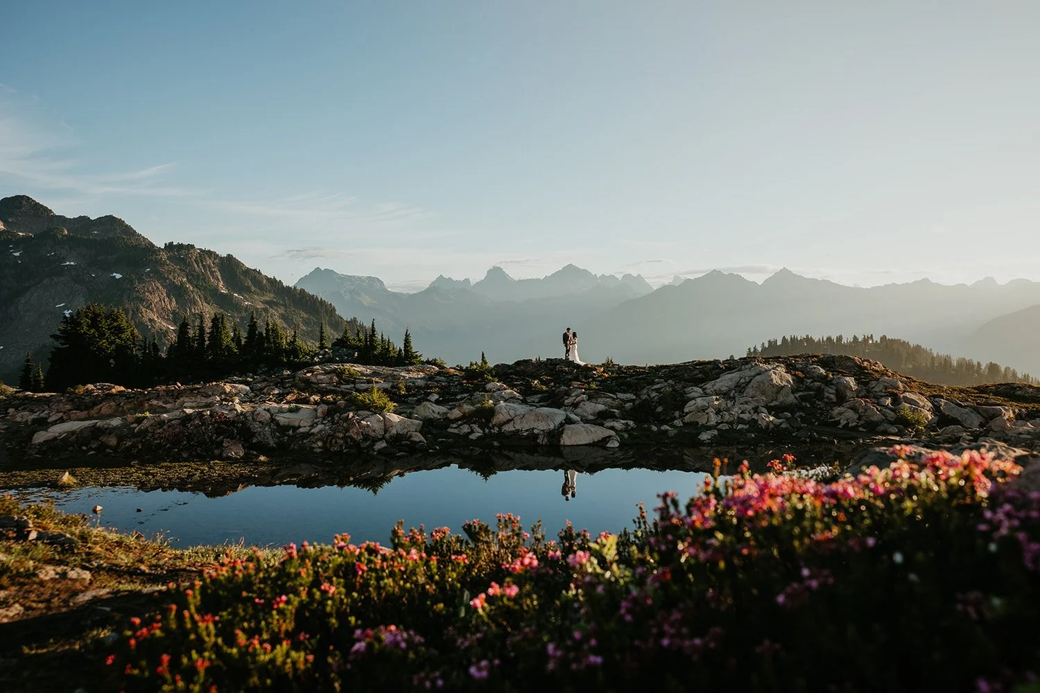 An eloping couple stands beyond a high-elevation alpine lake in the North Cascades surrounded by rocky peaks and summer wildflowers