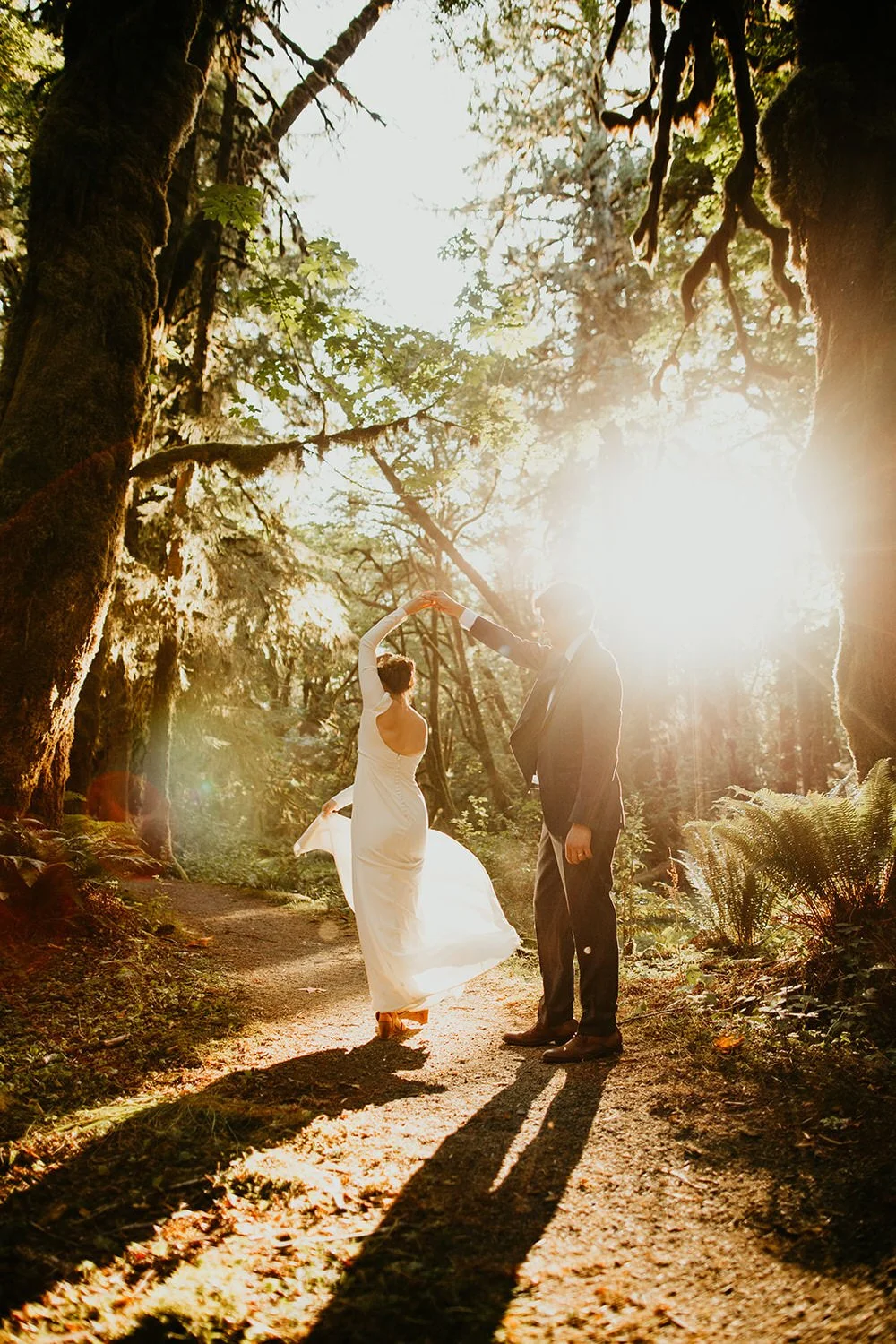 Couple twirling together on a wooded path as sunlight streams through the trees, casting long shadows across the trail.