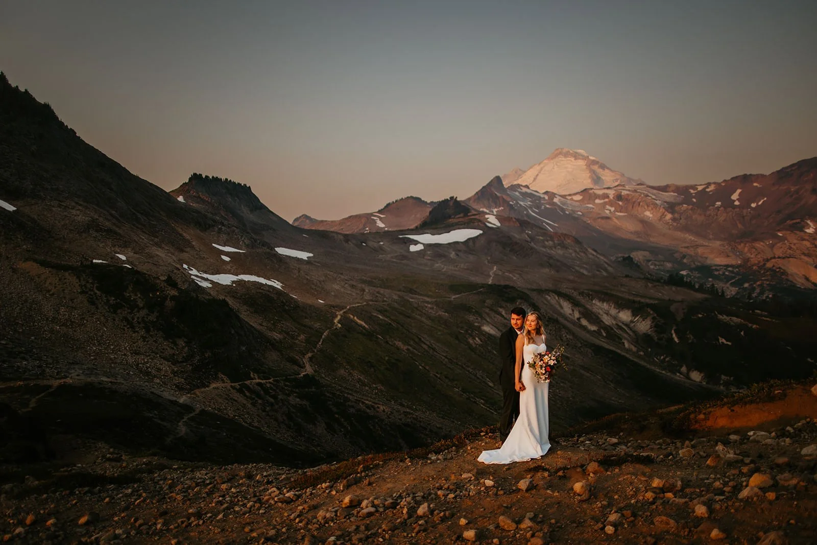Newlyweds standing on an alpine ridge at dusk with Mount Rainier in the distance, soft pink and amber tones settling over the Washington landscape.