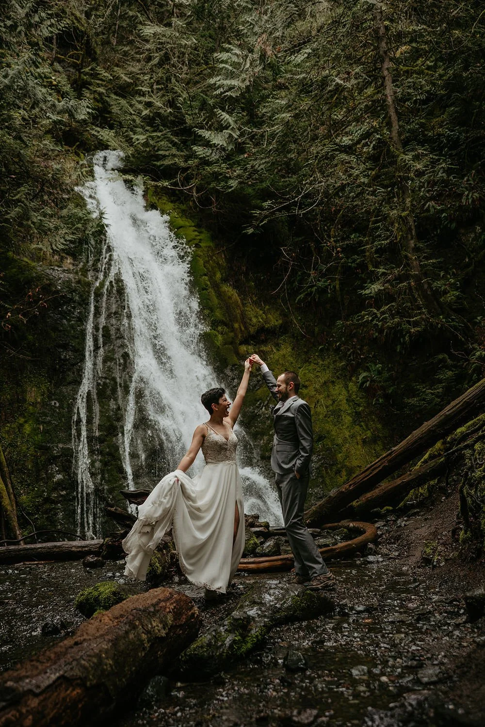Eloping couple dancing barefoot beside a forest waterfall surrounded by mossy rocks, fallen logs, and lush greenery