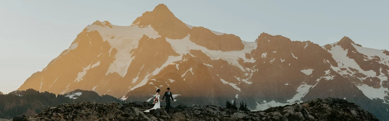A couple walks hand in hand across a rocky alpine ridge as warm evening light hits a dramatic, snow-dusted mountain face.