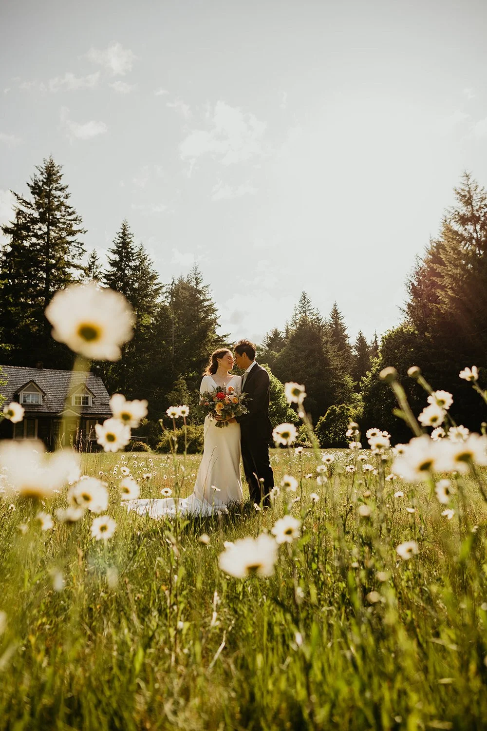 Eloping couple standing together in a field of wildflowers with evergreen trees and a rustic lodge in the background on a sunny summer day.