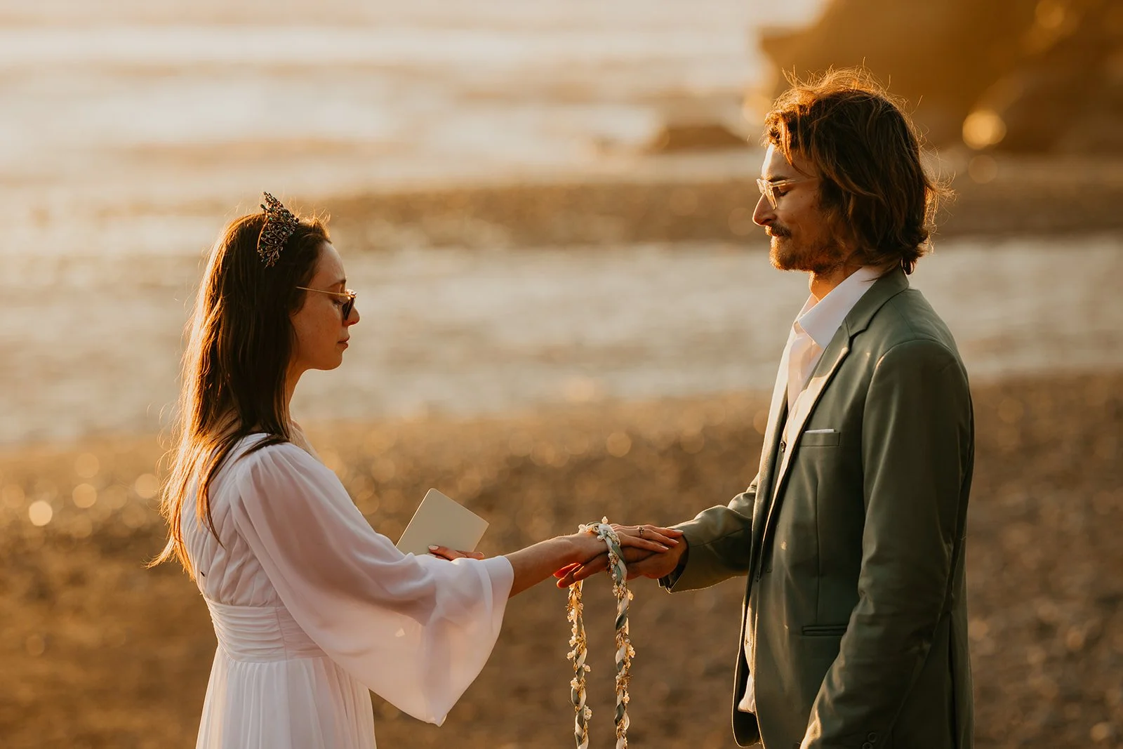 Couple exchanging vows on a rocky beach at golden hour with ocean waves behind them.