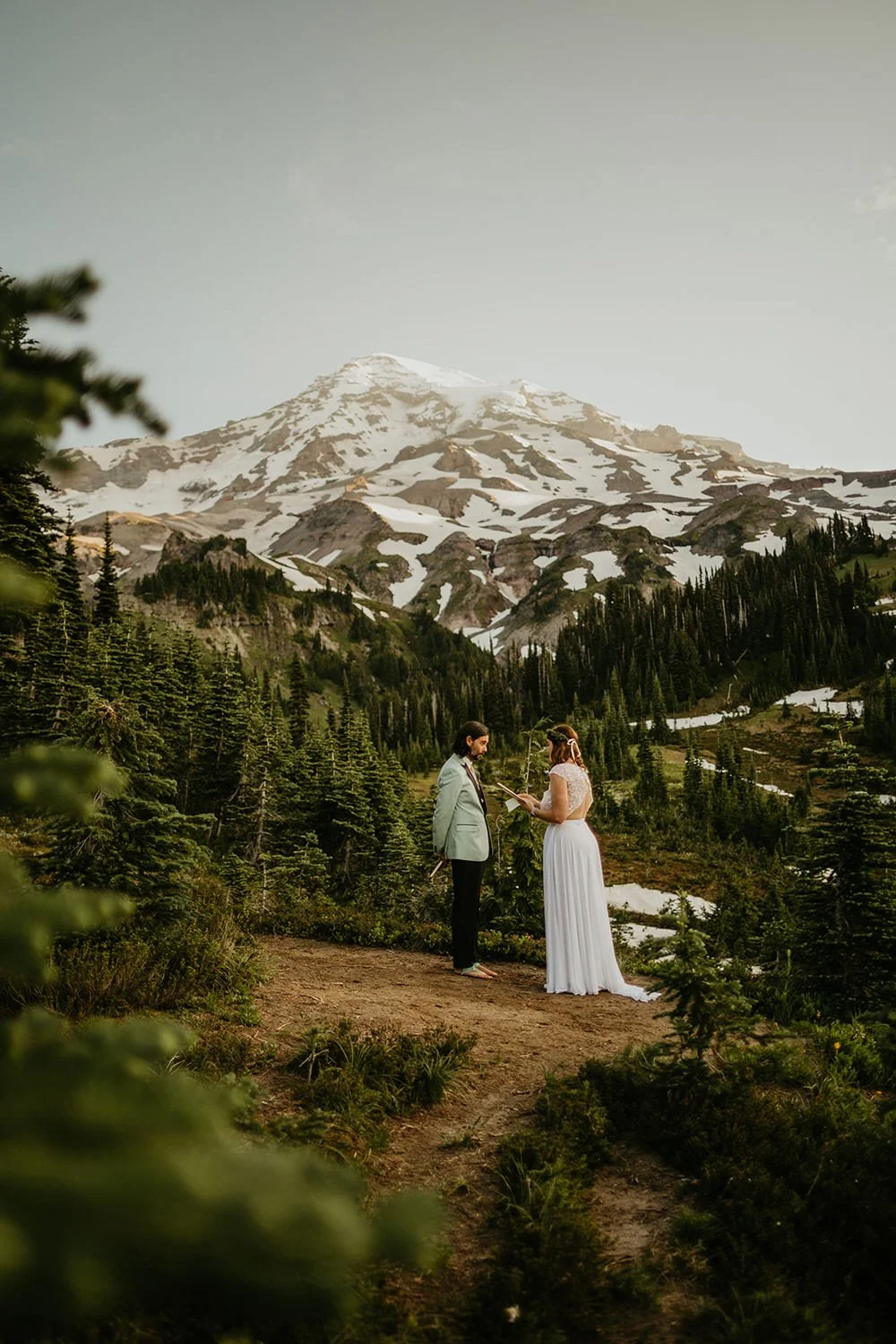 A couple stands facing each other in a forest clearing with Mount Rainier visible beyond evergreen trees and alpine terrain.